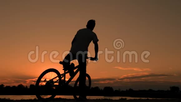 一个穿T恤的青少年坐在自行车上在黎明的清晨骑着自行车穿过河流或湖泊骑自行车的人的剪影视频的预览图