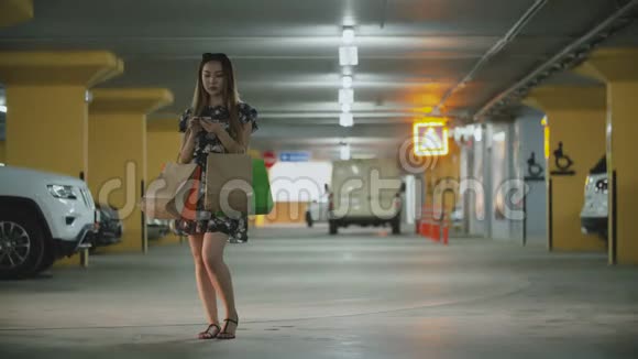 一个带购物袋的亚洲女人站在停车场用智能手机叫出租车视频的预览图