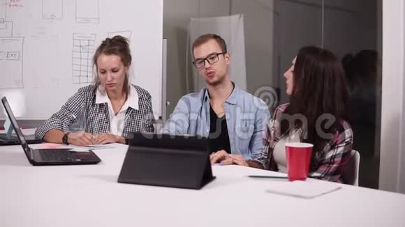 戴眼镜的年轻人和两个随意坐在办公桌前的女人桌子上放着笔记本电脑和平板电脑创意十足视频的预览图