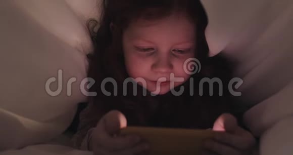 红头发的小女孩晚上在黄色智能手机上看视频她躲在毯子下面视频的预览图