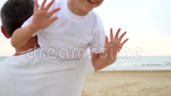 父子俩在沙滩上玩耍一个男人扛着他的肩膀一个快乐的笑着的男孩挥动着他的手视频的预览图