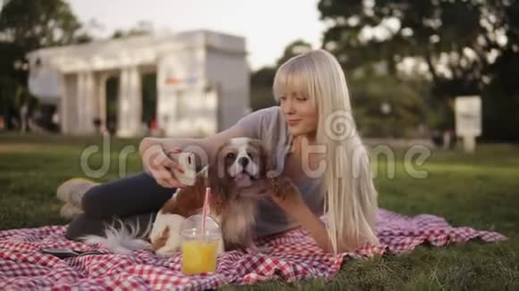 一位金发碧眼的女人躺在公园的格子布上试图用她的猎犬拍一张自拍照片视频的预览图