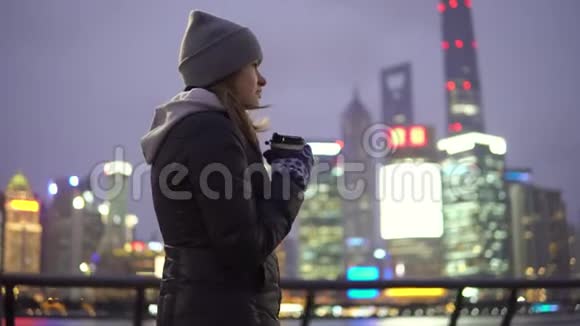 一个穿着黑色夹克和帽子的年轻女孩手里拿着咖啡在美丽的上海走来走去视频的预览图