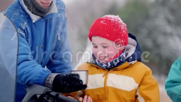 冬季森林一家年轻的家庭穿着五颜六色的衣服在雪地摩托车旁休息在一个杯子里喝着热饮料视频的预览图