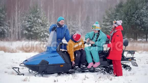 冬季森林一家年轻的家庭穿着五颜六色的衣服在雪地摩托车旁休息喝热饮料视频的预览图