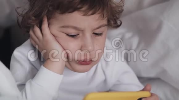带黄色智能手机的有趣的小男孩躺在床上用智能手机他在电话里看视频视频的预览图