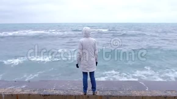 穿着白色夹克的女人站在海滩上双臂张开看着暴风雨的波浪后景视频的预览图