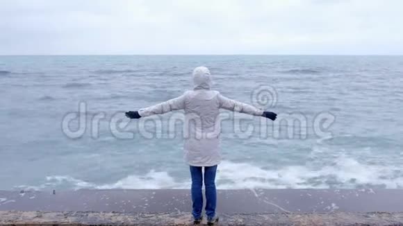 穿着白色夹克的女人站在海滩上双臂张开看着暴风雨的波浪后景视频的预览图