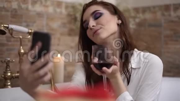 年轻女子带着鲜艳的妆容自拍坐在浴缸里用红酒杯可爱的女孩在浴室里放松视频的预览图