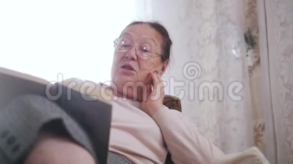 一位老太太喜欢坐在椅子上在窗户的背景上大声朗读闭合视频的预览图