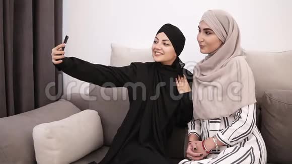 两个阿拉伯女人戴着头巾坐在灰色沙发上在家一起自拍白色的墙上视频的预览图