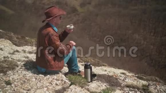 山上坐着一个男人戴着牛仔帽皮夹克蓝色牛仔裤和眼镜一个人喝热水瓶里的茶视频的预览图