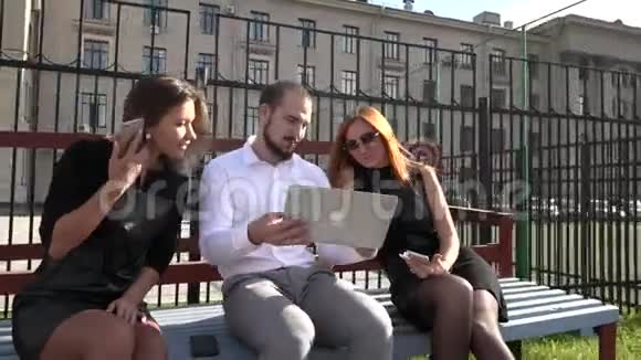 两个女生和男生在阳光明媚的天气里用笔记本电脑在长凳上交流视频的预览图