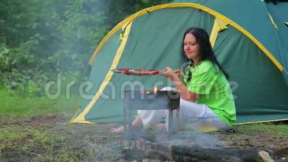一个穿着绿色T恤的女人坐在一个帐篷的背景下的森林里在她的胸罩上准备晚餐视频的预览图