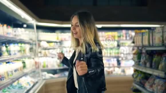 在超市里穿着黑色夹克的快乐年轻女人在货架上的商品和奶制品中跳舞旋转视频的预览图