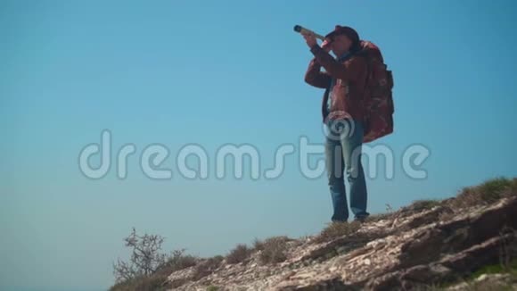 戴牛仔帽皮夹克蓝色牛仔裤肩上背着旅游背包的人一个人透过望远镜看视频的预览图