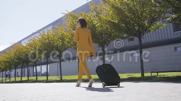 一位穿着正装的年轻迷人的女商人拉着手提箱用智能手机聊天匆匆忙忙地去做生意视频的预览图