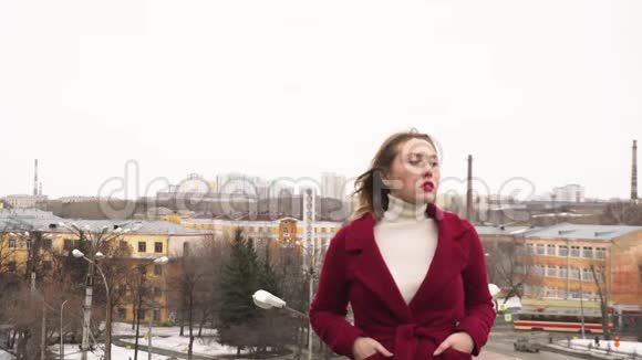穿着红色外套和白色高领毛衣的年轻漂亮女人的特写站在屋顶上看着远处媒体照片视频的预览图