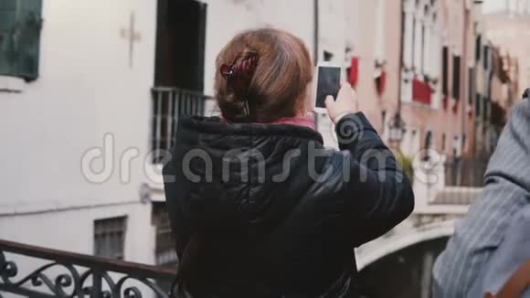 背景快乐资深女游客在意大利威尼斯美丽的秋水渠街拍摄智能手机照片视频的预览图