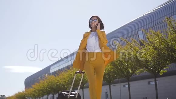 穿着西装戴着太阳镜的商务女性在火车站附近带着手提行李走路时用智能手机聊天或者视频的预览图