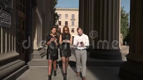 英俊的男学生和两名女学生靠近大学用智能手机相互交流视频的预览图