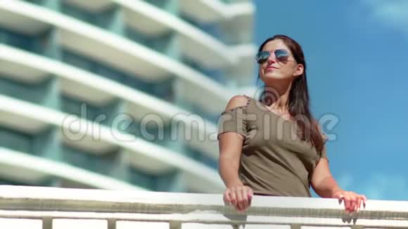 迷人的微笑晒黑的女人戴着太阳镜在豪华的现代酒店阳台上放松视频的预览图