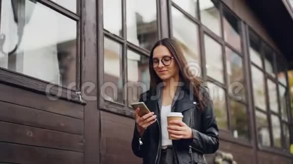 戴眼镜的可爱年轻女子用智能手机微笑着走在街上拿着外卖的慢动作视频的预览图