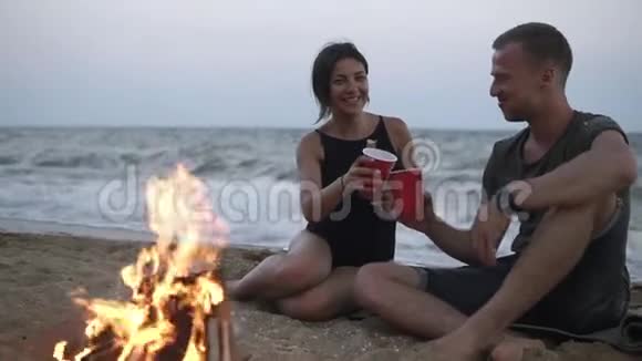 美丽的白种夫妇在篝火旁的海滩上享受时光喝着红色的杯子穿黑色衣服的女人视频的预览图