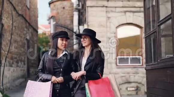 穿着时髦服装的快乐女孩手挽手地走在街上手提购物袋谈论社交活动视频的预览图