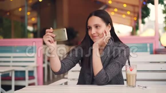 一位优雅的年轻女士正在用智能手机打电话在露天咖啡厅放松边喝奶昔边聊天视频的预览图
