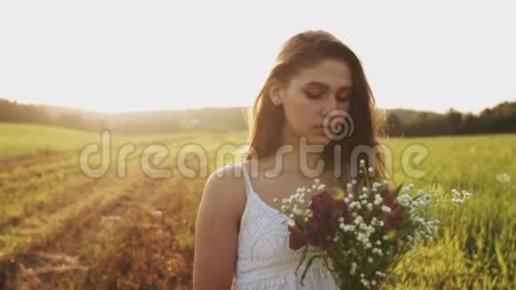 夏日傍晚日落时分站在草地上的女孩手捧着一束野花看着镜头视频的预览图