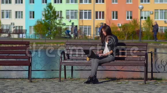 公园里的一个女孩坐在湖边的长凳上拿着笔记本电脑喝着咖啡视频的预览图