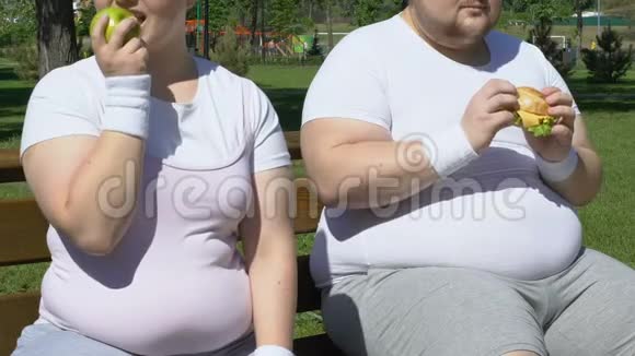 快乐的胖女孩在锻炼后吃苹果嘲笑有汉堡的肥胖男人视频的预览图