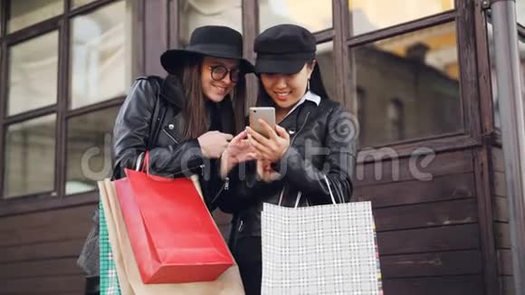 兴奋的女孩们正在使用智能手机触摸屏幕站在街上在秋日拿着购物袋视频的预览图