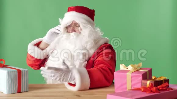 满意的圣诞老人在电话里说话笑着拿着礼物坐在桌子旁背景是绿色的视频的预览图