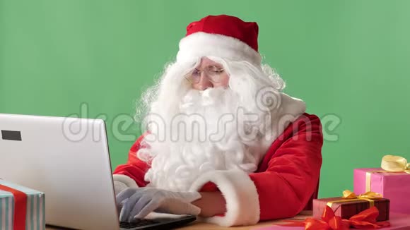 严肃的圣诞老人坐在笔记本电脑上打字看着相机挥动着他的手打招呼的手势是彩色的视频的预览图