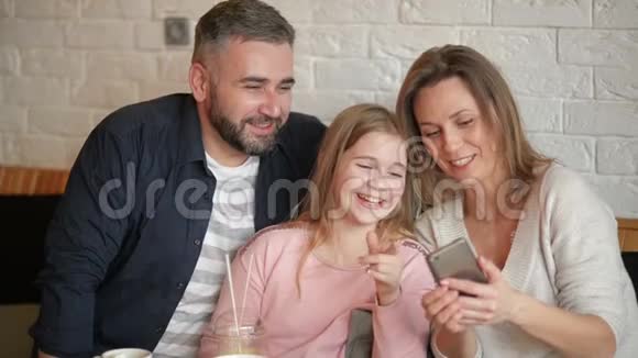 快乐的母亲父亲和小女孩有乐趣并通过智能手机在咖啡厅视频的预览图