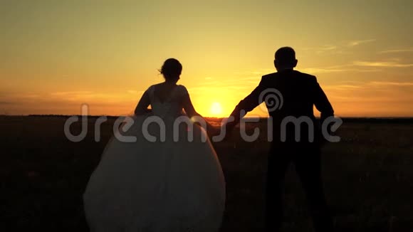 快乐的情侣旅行新娘和新郎在夕阳的照耀下跑过田野恋人们携手在草地上视频的预览图