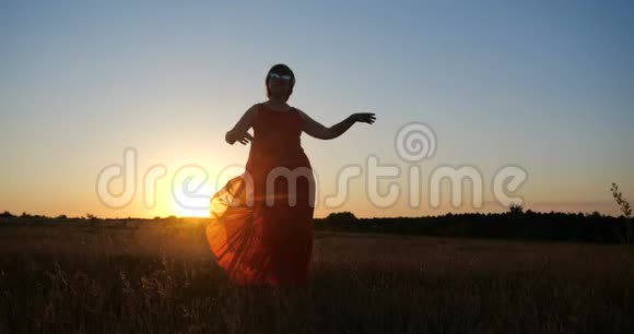 在夕阳下一个穿着红色裙子在无边无际的田野里跳舞的受启发的女人视频的预览图