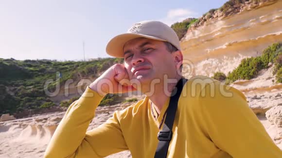 一个穿着黄色毛衣和帽子的疲惫的中年男人坐在岩石绿色灌木和石头的背景下视频的预览图