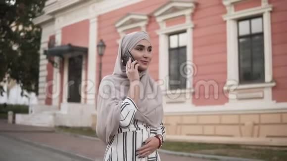 戴着头巾化妆的阿拉伯穆斯林妇女走在街上在美丽的老人背景下打电话视频的预览图