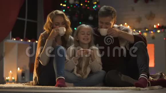 一家人坐在地板上喝热茶圣诞树和闪闪发光的灯视频的预览图