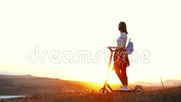一个骑着电动滑板车的女孩站着背对着夕阳高兴地抬起手慢悠悠地视频的预览图