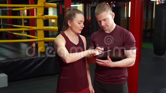 肌肉型个人教练在剪贴板上写作并在交叉训练健身房向年轻女性讨论训练计划视频的预览图