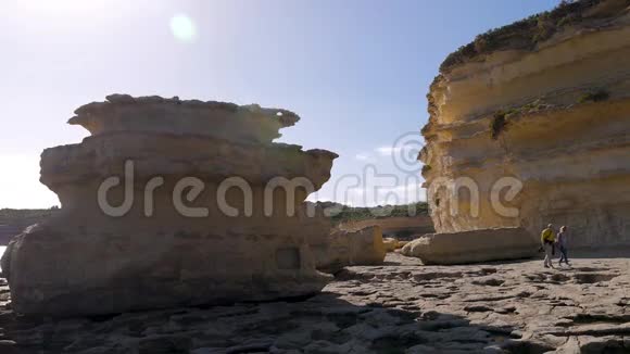 在一个风和日丽的日子里两个游客沿着海洋的岩石海岸在巨大的棕色石头粘土中散步家庭视频的预览图