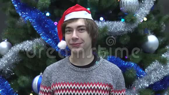 圣诞帽中微笑的年轻人的肖像看圣诞树背景上的相机视频的预览图