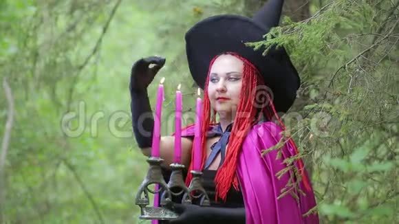 年轻的女巫红发黑帽子和覆盆子斗篷在森林里用蜡烛变魔术视频的预览图