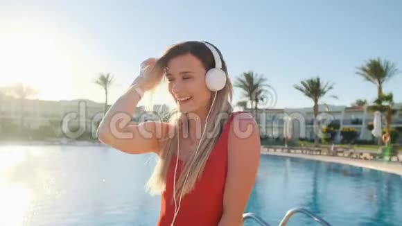 美丽的女士穿着红色泳衣和耳机在清澈的蓝色游泳池背景下跳舞玩得开心听得开心视频的预览图