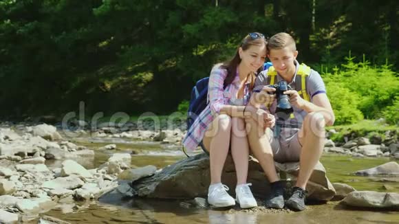 年轻的游客看着相机上拍摄的照片他们坐在靠近山河和森林的岩石上视频的预览图