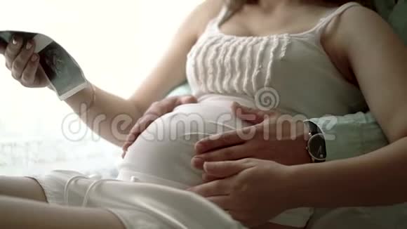 一对夫妇看着沙发上婴儿的超声波扫描双手抱着肚子视频的预览图
